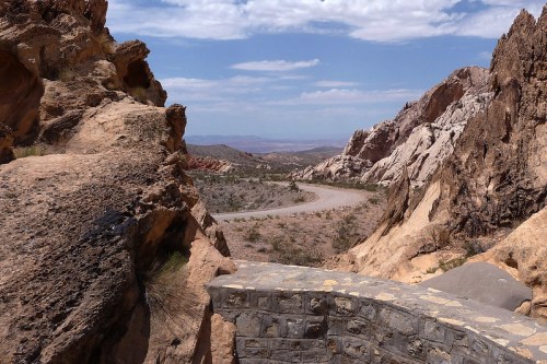 A winding road cuts through rocky terrain under a blue sky with scattered clouds. Desert landscape in the background.