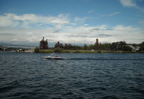 A boat glides across a river with industrial buildings and a bridge in the background under a cloudy sky.