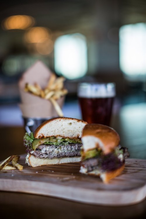 A close-up of a juicy burger with lettuce and pickles, served on a wooden board with fries and a glass of beer in the background.