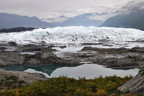A vast glacier reflects in a calm lake, surrounded by rocky terrain and distant mountains under a cloudy sky.
