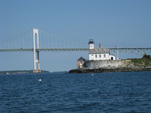A lighthouse and historic building by the water, with a large bridge spanning the background under a clear blue sky.