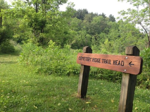 Wooden sign for Cemetery Ridge Trail Head, surrounded by lush greenery and trees in a peaceful outdoor setting.