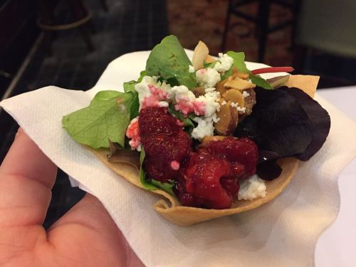 A hand holding a small salad in a tortilla cup, featuring greens, cheese, nuts, and raspberries.