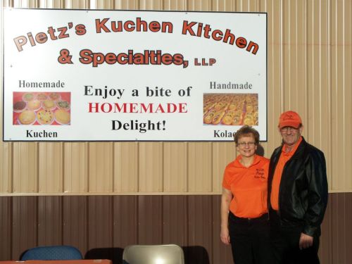 A couple stands in front of a sign for Pletz's Kuchen Kitchen, promoting homemade kuchen and kolaches.