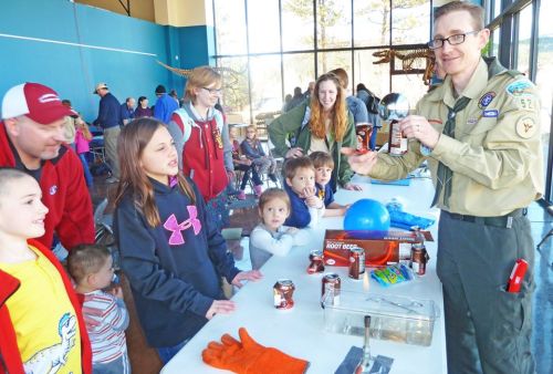 A group of children and adults gather around a table as a man in a uniform demonstrates an experiment with soda cans.