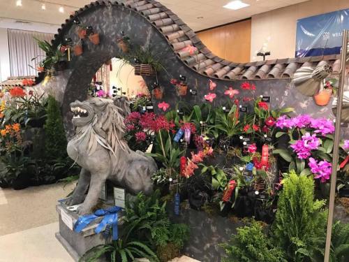 A decorative display featuring a stone lion and vibrant orchids, with ribbons, in a floral exhibition setting.
