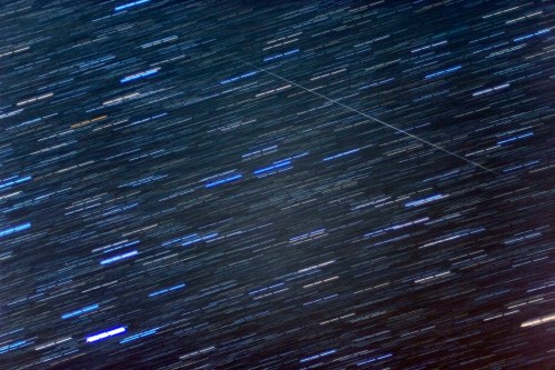 A blurred night sky filled with streaks of stars and a faint line, possibly a satellite or meteor, cutting through the scene.
