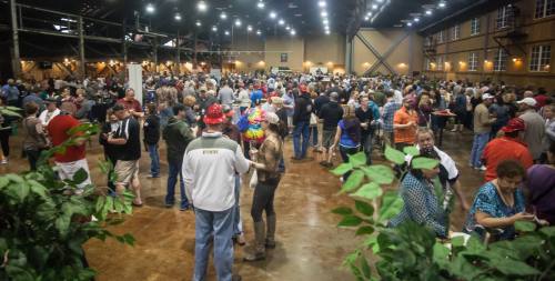 A large crowd of people socializing in a spacious indoor venue with greenery in the foreground.