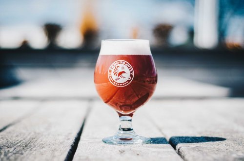 A glass of amber beer with a logo sits on a wooden surface, with a blurred outdoor background.