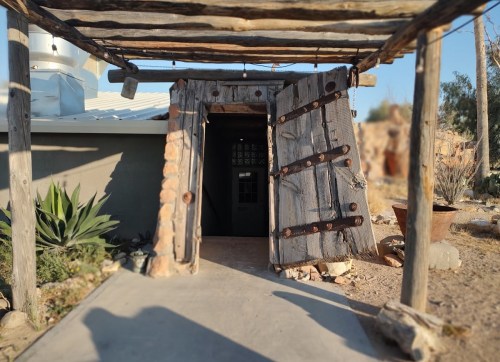 A rustic wooden door with metal accents leads into a stone structure, surrounded by desert plants and rocky terrain.