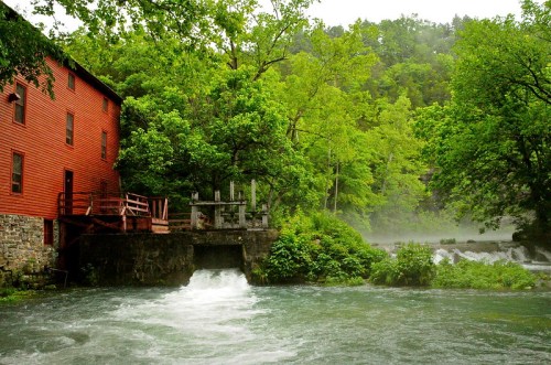 A red mill building beside a rushing river, surrounded by lush green trees and misty water.