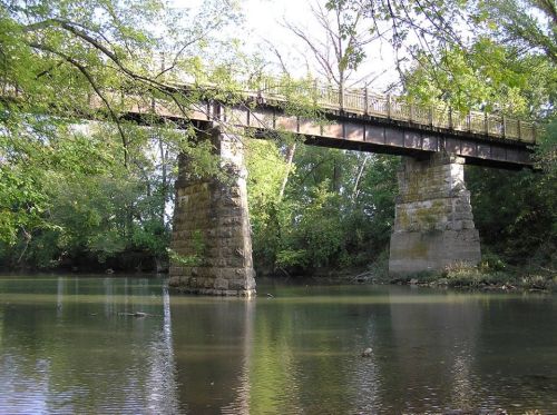 A sturdy bridge spans a calm river, surrounded by lush green trees and reflections on the water's surface.