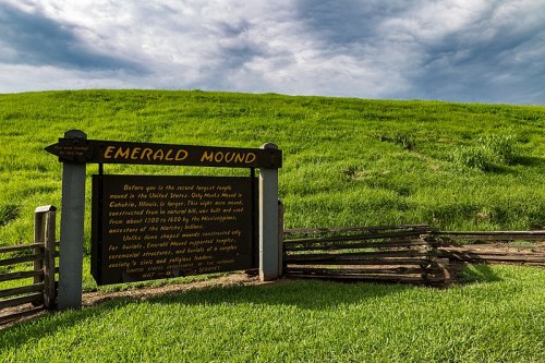 Sign for Emerald Mound, a large Native American earthwork, with a green grassy mound and cloudy sky in the background.