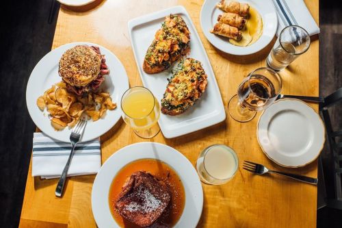 A table set with a burger, chips, two baked dishes, spring rolls, and drinks, showcasing a variety of delicious foods.