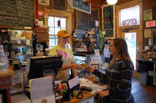 A barista hands a pastry to a customer at a cozy café with a chalkboard menu and colorful decor.