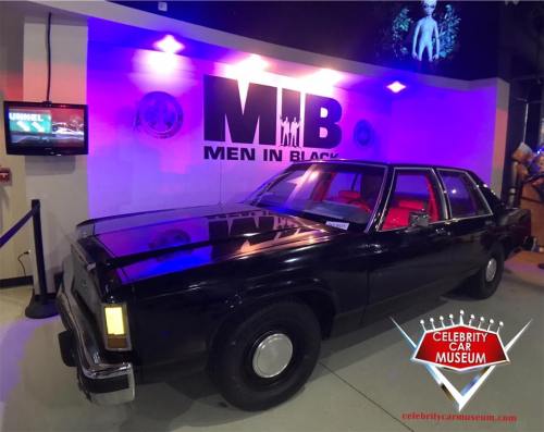 A black car displayed in a museum, with "MIB" and "Men in Black" signage in the background.