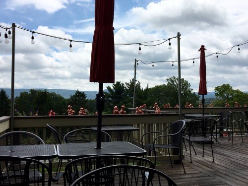 A scenic outdoor deck with tables, umbrellas, and string lights, overlooking a cloudy landscape with trees in the distance.