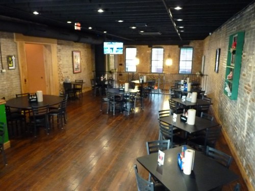 Interior of a restaurant with wooden floors, black tables, and chairs, featuring exposed brick walls and a TV.