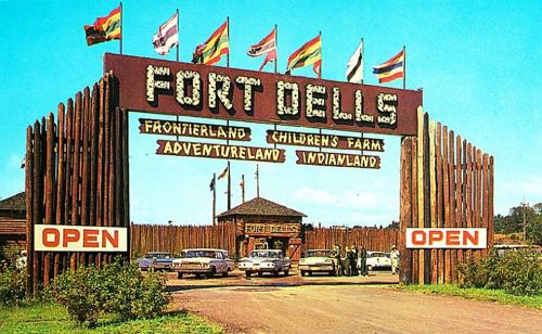 Vintage entrance sign for Fort Dells, featuring flags and wooden structures, with cars parked in front.
