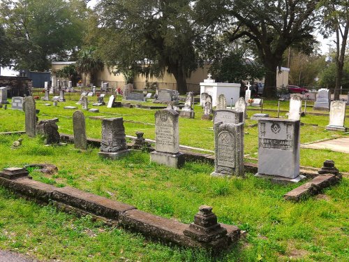 A cemetery with various gravestones and monuments surrounded by grass and trees under a clear sky.