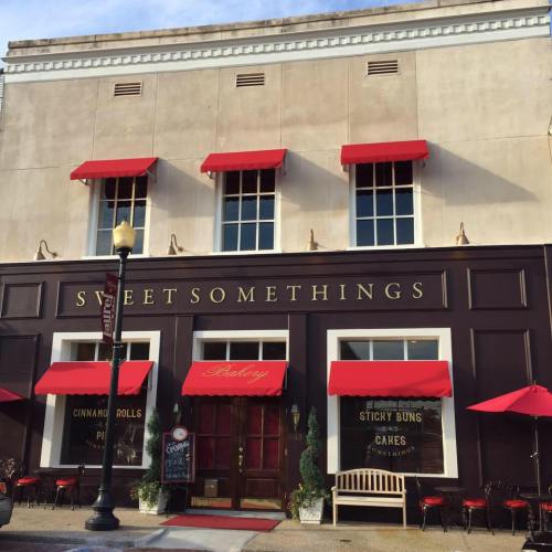 A charming bakery with red awnings, featuring a sign that reads "Sweet Somethings" and outdoor seating.