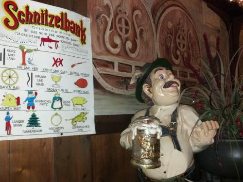 A wooden figure holding a beer stein stands next to a colorful Schnitzelbank sign in a rustic setting.