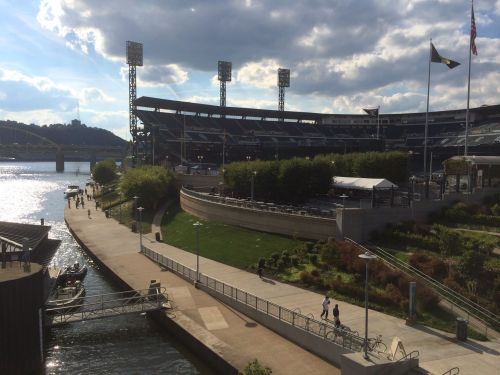 A scenic view of a waterfront park near a baseball stadium, with people walking along the path and boats on the water.