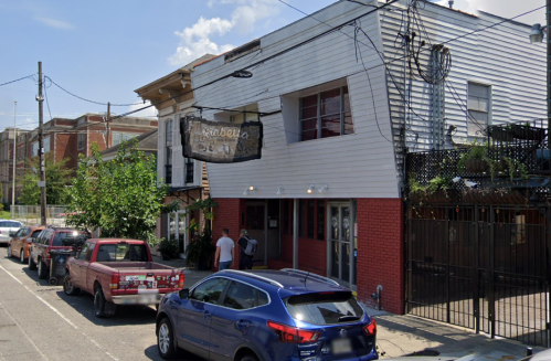 A street view of a restaurant with a sign, surrounded by parked cars and greenery on a sunny day.