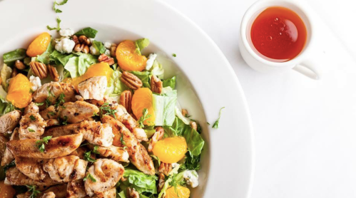 A fresh salad with grilled chicken, mandarin oranges, pecans, and feta cheese, served with a small cup of dressing.