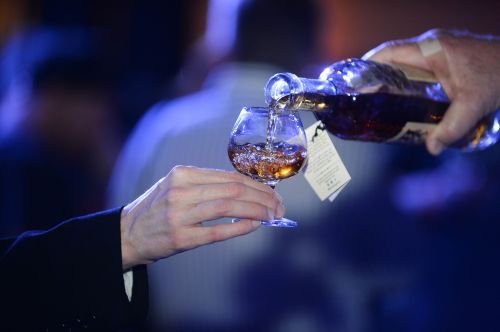 A hand pours a drink into a glass, with a blurred background of people and soft lighting.
