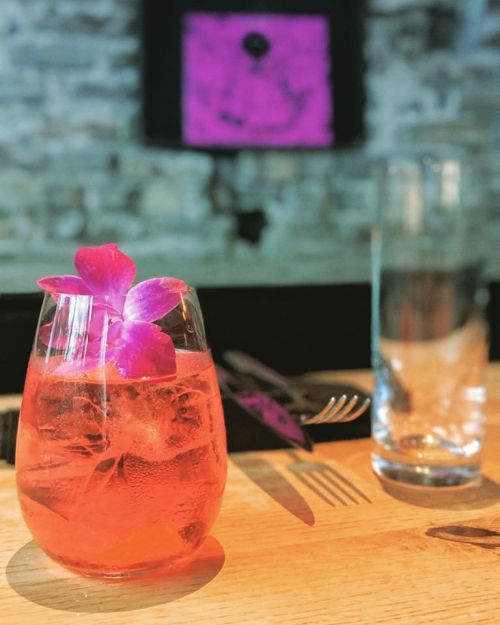 A vibrant pink cocktail garnished with a purple flower, placed on a wooden table with a glass of water in the background.