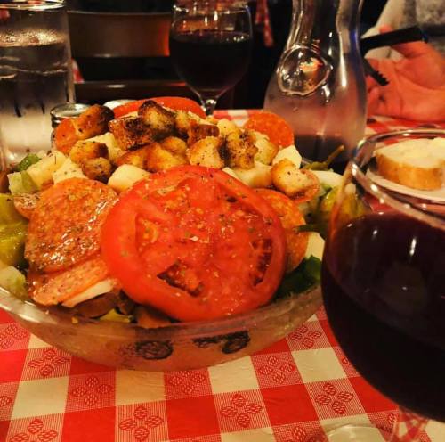 A colorful salad with croutons, tomatoes, and pepperoni, accompanied by glasses of red wine on a checkered tablecloth.