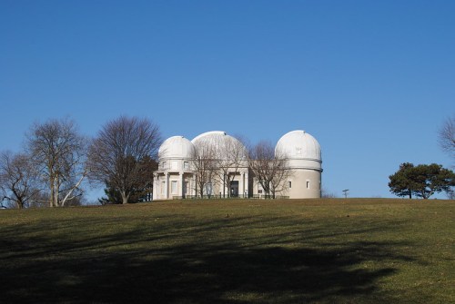 A large observatory with three domes sits atop a grassy hill under a clear blue sky, surrounded by bare trees.