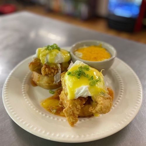 A plate with two servings of fried chicken and poached eggs on biscuits, topped with hollandaise sauce and green onions, plus a side of cheese grits.