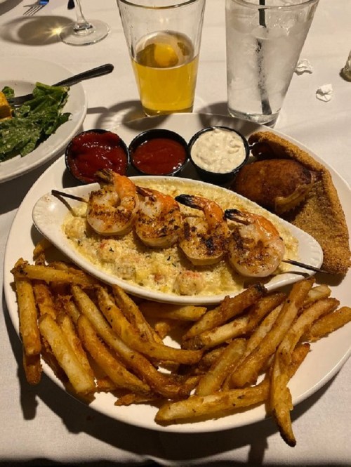 A plate of shrimp skewers, fries, and dipping sauces, with a salad and drinks in the background.