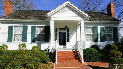 A charming white house with green shutters, a front porch, and brick steps surrounded by neatly trimmed bushes.