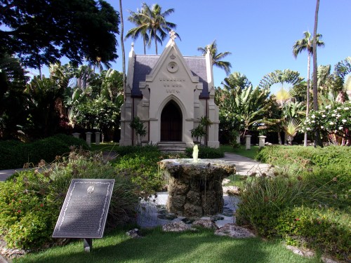 A small, ornate chapel surrounded by lush greenery and palm trees, with a fountain and informational plaque in the foreground.