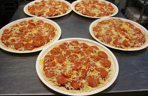 Five pizza trays topped with pepperoni and shredded cheese, ready for baking.