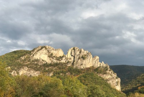 A rugged mountain peak rises against a cloudy sky, surrounded by lush green trees and hills.