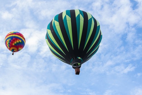 Two colorful hot air balloons float against a blue sky with scattered clouds. One has a striped green design, the other is multicolored.