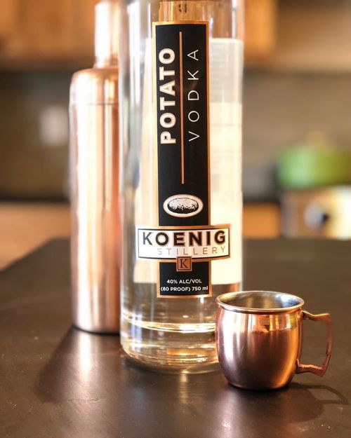 A bottle of Koenig Potato Vodka next to a copper shot glass and a cocktail shaker on a dark surface.