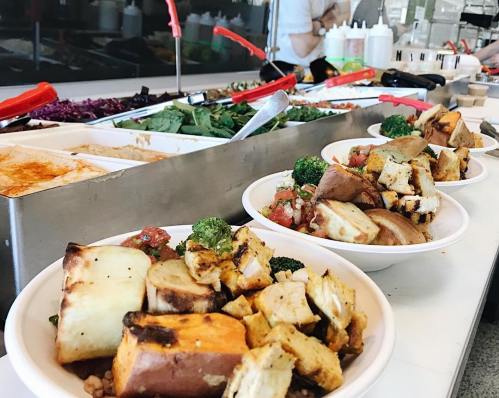 Bowls of colorful food with grilled vegetables and toppings, set against a backdrop of a salad bar.