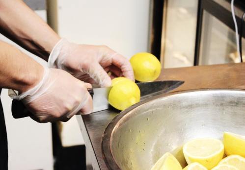 A person wearing gloves slices lemons on a countertop next to a bowl of lemon halves.