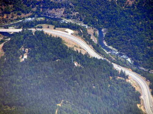 Aerial view of winding roads through lush green forests and a river, surrounded by mountainous terrain.