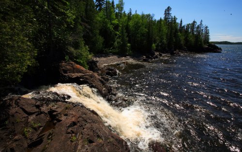 A serene riverside scene with a waterfall cascading over rocks, surrounded by lush green trees and a calm blue lake.
