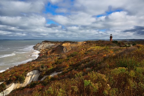 A coastal landscape featuring cliffs, a lighthouse, and colorful autumn foliage under a cloudy sky.
