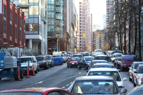Busy city street filled with parked and moving cars, surrounded by modern buildings and trees.