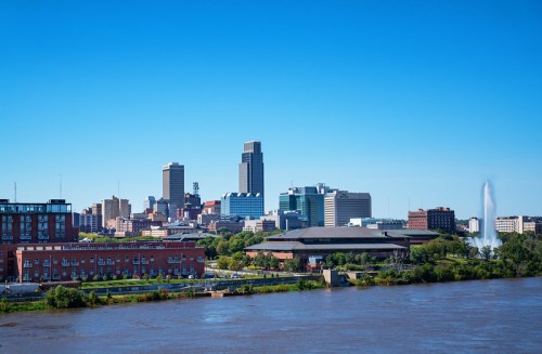 A clear blue sky over a city skyline with tall buildings, a river in the foreground, and a fountain in the background.