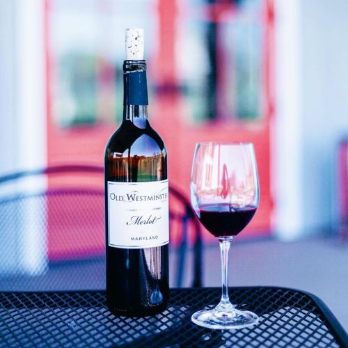 A bottle of Old Westminster Merlot next to a glass of red wine on a table, with a blurred red background.