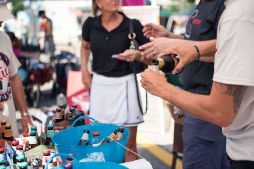 A person pours a drink from a bottle at a market stall, surrounded by others and various beverage containers.